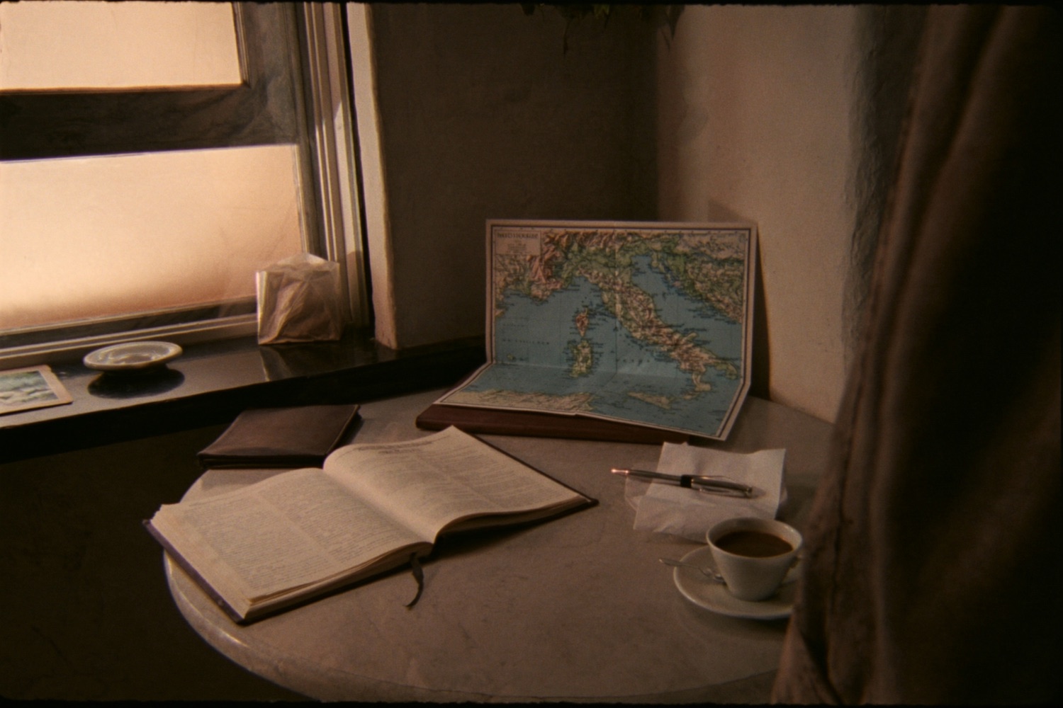 A sunlit window table with a map of Italy, an open book, a journal and an espresso